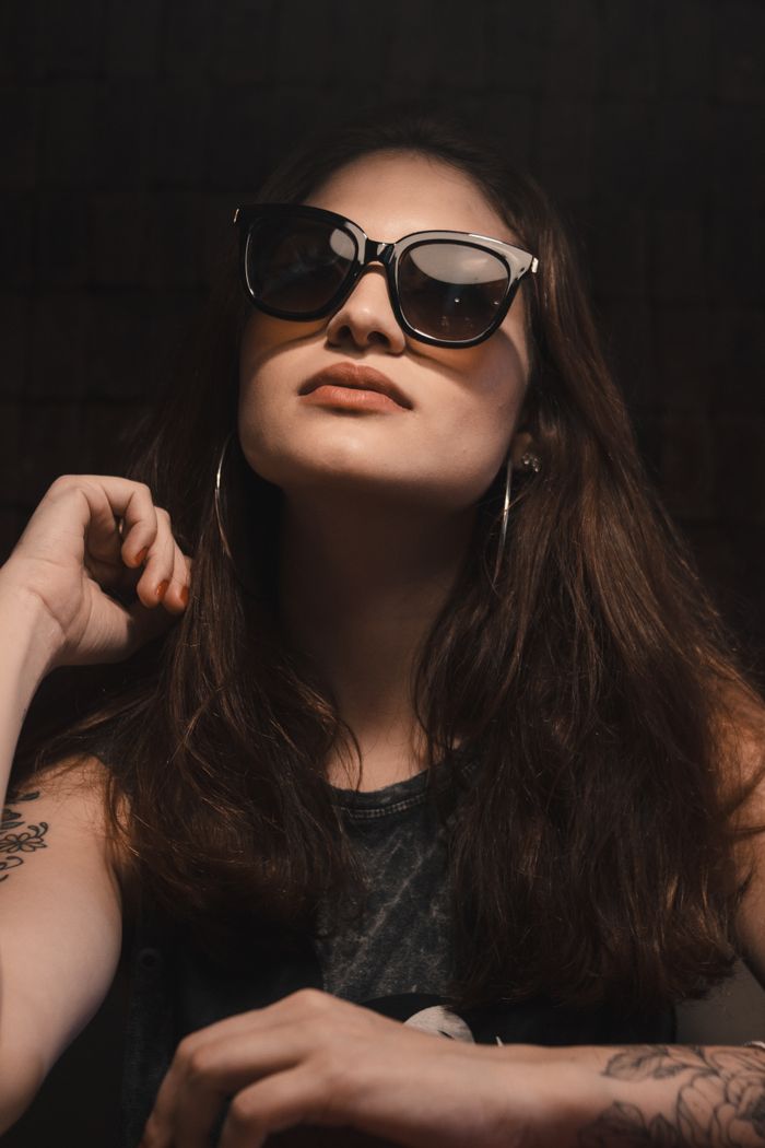 A fashionable woman with long hair poses confidently wearing sunglasses, showing her tattoos indoors.