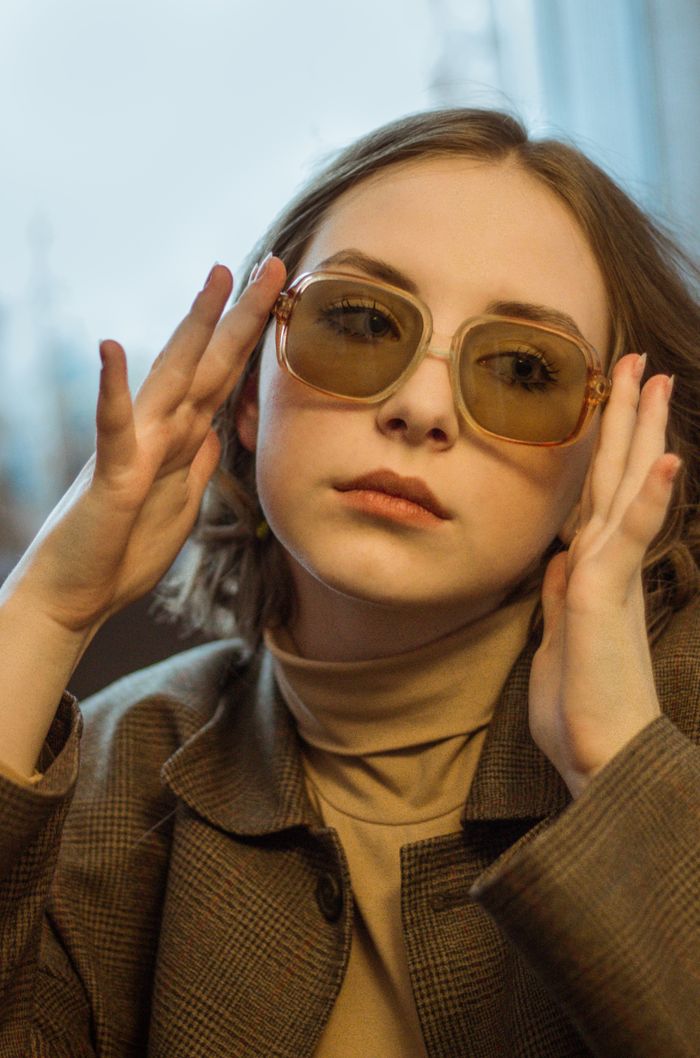 Young woman adjusting fashionable sunglasses indoors, showcasing modern eyewear style.